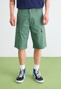 Green cargo shorts with side pockets, made from textured fabric. Worn with high-top black and blue sneakers and white socks.