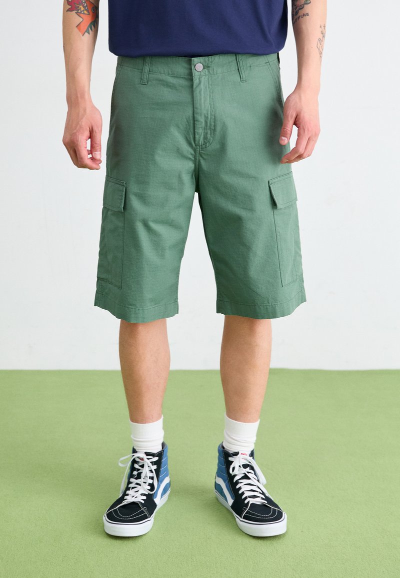 Green cargo shorts with side pockets, made from textured fabric. Worn with high-top black and blue sneakers and white socks.
