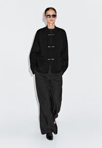 Vrouw draagt een oversized zwarte jas met voorste sluitingen, brede zwarte broek, zwarte laarzen en donkere zonnebril, staand tegen een effen witte achtergrond.