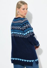 Mörkblå tröja med ett geometriskt Fair Isle-mönster i ljusblått och vitt. Långa ärmar, avslappnad passform, sidoflikar vid nederkanten.