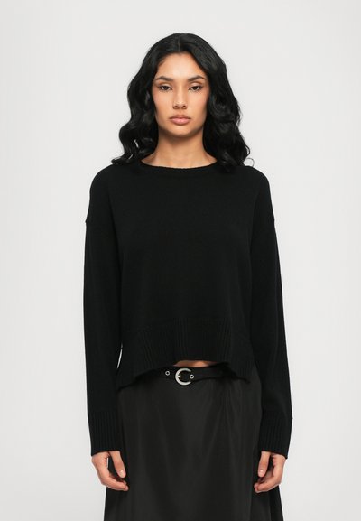 Pull en tricot noir avec un col rond, des épaules tombantes et un ourlet côtelé. Porté sur une jupe noire avec une ceinture à boucle argentée.
