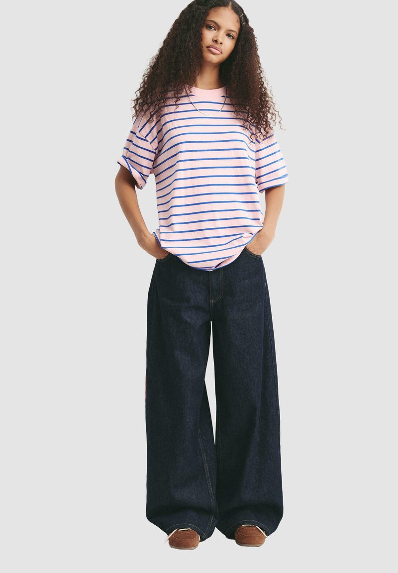 Rózsaszín csíkos, rövid ujjú ing és széles szárú sötét denim nadrág. Az ing kék csíkokkal rendelkezik; a nadrág laza fitneszben készült, oldalsó zsebekkel.