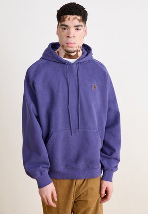 Sudadera morada con bolsillo canguro y cordón. Material de algodón con un parche de logo en el pecho. El modelo lleva pantalones de pana.