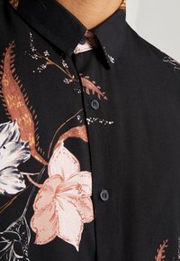 Chemise noire à motifs floraux avec des lys roses pâles, col boutonné et boutons bleus, mettant en valeur une surface en tissu lisse et un design détaillé.