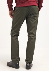 Une personne portant un pantalon vert olive, des baskets noires et une chemise rouge à manches longues, se tenant avec les mains près des poches, de dos.