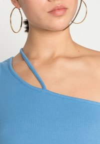 Top azul-claro canelado com uma alça, apresentando um decote limpo e uma textura subtil. Modelo usa grandes brincos de argola dourados.