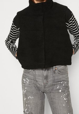 Person iført en sort, fluffy vest over en sort og hvid stribet langærmet skjorte og grå jeans med hvide malingstænk.