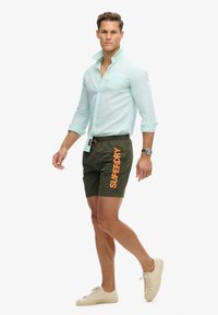 Mann, der ein hellblaues Hemd mit Knöpfen, dunkelgrüne Shorts mit dem orangefarbenen Schriftzug "Superdry", weiße Sneakers und eine Armbanduhr am linken Handgelenk trägt.