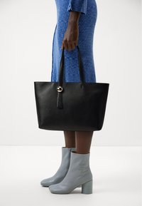 Sac en cuir noir avec deux poignées, mettant en valeur un accent doré circulaire. Associé à des bottines bleu clair avec un talon carré.