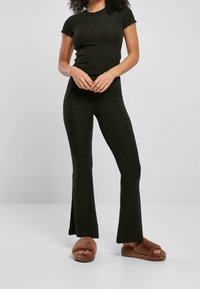 Vrouw in een zwarte ribgebreide top met korte mouwen en bijpassende uitlopende broek, met bruine fuzzy slippers, staande tegen een effen grijze achtergrond.
