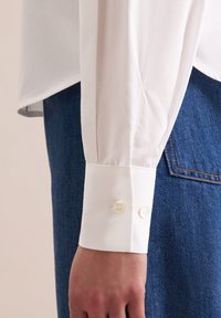 Camicia bianca in cotone con maniche strutturate, dotata di due bottoni di chiusura al polsino; abbinata a un denim blu scuro. Texture liscia.