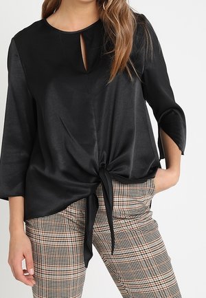 Femme portant un chemisier noir à manches longues avec un nœud devant et un décolleté en forme de serrure, associé à un pantalon à carreaux marron et beige.