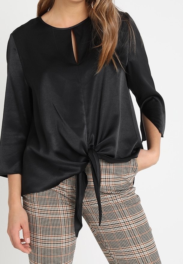 Femme portant un chemisier noir à manches longues avec un nœud devant et un décolleté en forme de serrure, associé à un pantalon à carreaux marron et beige.
