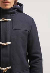 Abrigo de lana azul marino tipo duffle con capucha, que presenta cierres de toggles de madera y detalles de cuerda anudada, superficie texturizada y detalle en el bolsillo del pecho.