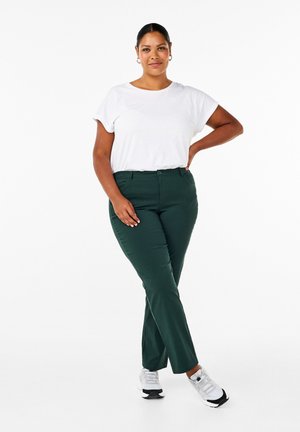 Gröna skräddarsydda byxor med en mjuk textur, kombinerat med en åtsittande vit T-shirt. Modell som bär vita och svarta sneakers, med händerna på höfterna.