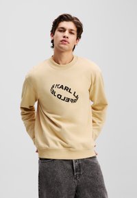 Sweatshirt beige à col ras du cou avec un texte noir imprimé en forme d'oval déformé, associé à un jean gris. Texture lisse et coupe décontractée.