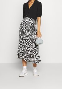 Blouse noire boutonnée associée à une jupe mi-longue imprimé zèbre noir et blanc, des baskets blanches et un petit sac à main bleu clair.