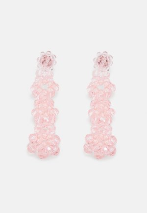 Pár visících náušnic z shluků průhledných růžových korálků vytvářejících svislý květinový vzor na bílém pozadí.
