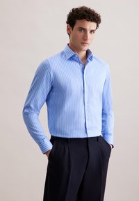 Camicia azzurro chiaro con strisce verticali bianche, maniche lunghe e colletto classico. Indossata con pantaloni scuri, mani nelle tasche.