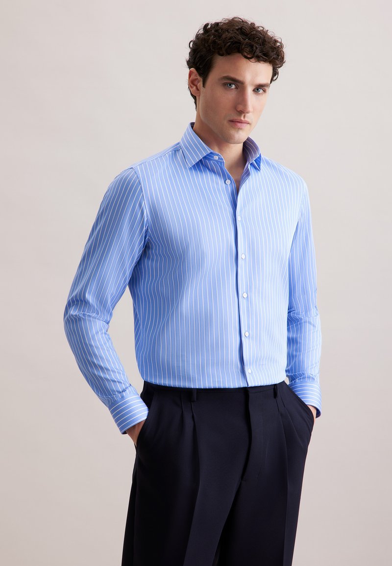 Camicia azzurro chiaro con strisce verticali bianche, maniche lunghe e colletto classico. Indossata con pantaloni scuri, mani nelle tasche.