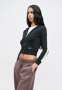 Moteris su ilgais juodais plaukais, vilkinti juodą trumpą užtrauktuko džinsinį švarką ir rožinės spalvos „Nike“ sportinius džinsus, stovinti su rankomis kišenėse prieš baltą foną.