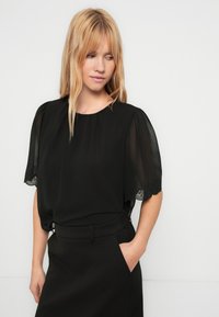 Schwarze Bluse mit transparenten, gepufften Ärmeln. Verfügt über einen Rundhalsausschnitt und Spitzenbesatz an den Ärmeln. Kombiniert mit maßgeschneiderten schwarzen Hosen.