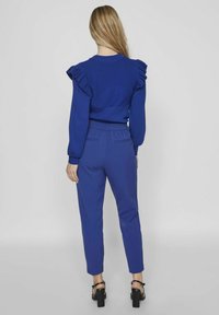 Ensemble bleu royal composé d'un pull côtelé avec des accents de manche à volants et d'un pantalon ajusté, tous deux en tissu lisse. Sandales à talons blocs noirs.