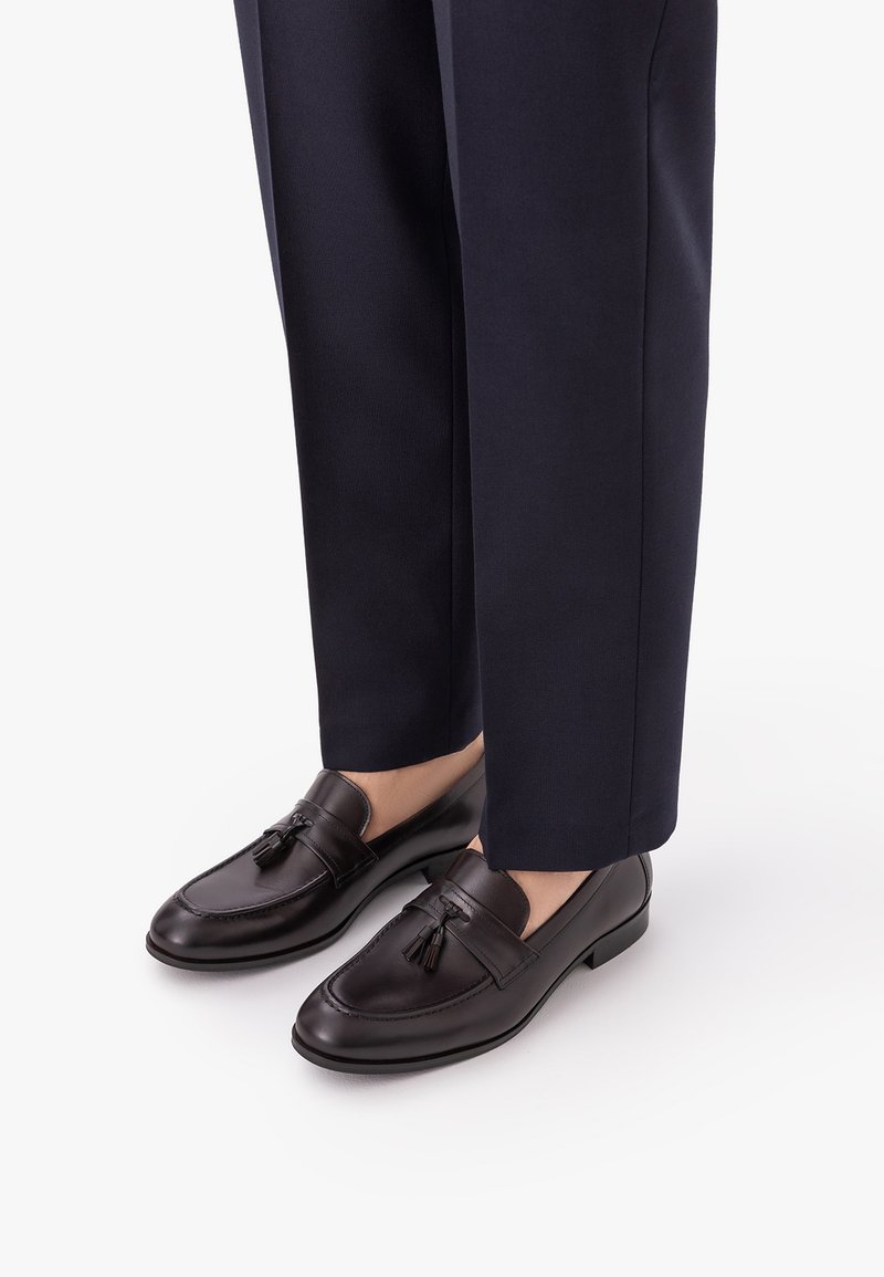 Σκούρα καφέ δερμάτινα loafers με φούντες, φορεμένα με σκούρο μπλε παντελόνι κοστουμιού, εμφανίζονται σε λευκό φόντο.