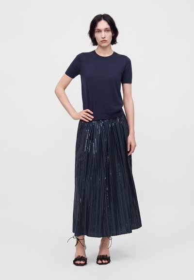 Femme portant un haut bleu marine à manches courtes et une longue jupe plissée brillante bleu marine, avec des talons noirs ouverts à l'avant, debout devant un fond blanc uni.