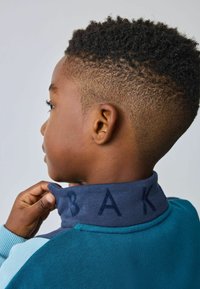 Chaqueta tipo pullover en verde azulado y azul con un alto collar de color navy que presenta "BAK" en letras texturizadas. Tejido de punto texturizado y corte relajado.