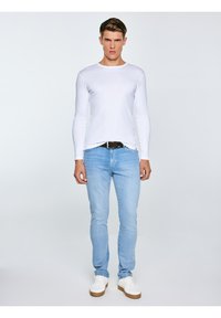 Jauns vīrietis stāv, valkājot baltu garroku kreklu, gaismazilas džinsas ar melnu jostu un baltas teniskas uz vienkrāsaina fona.