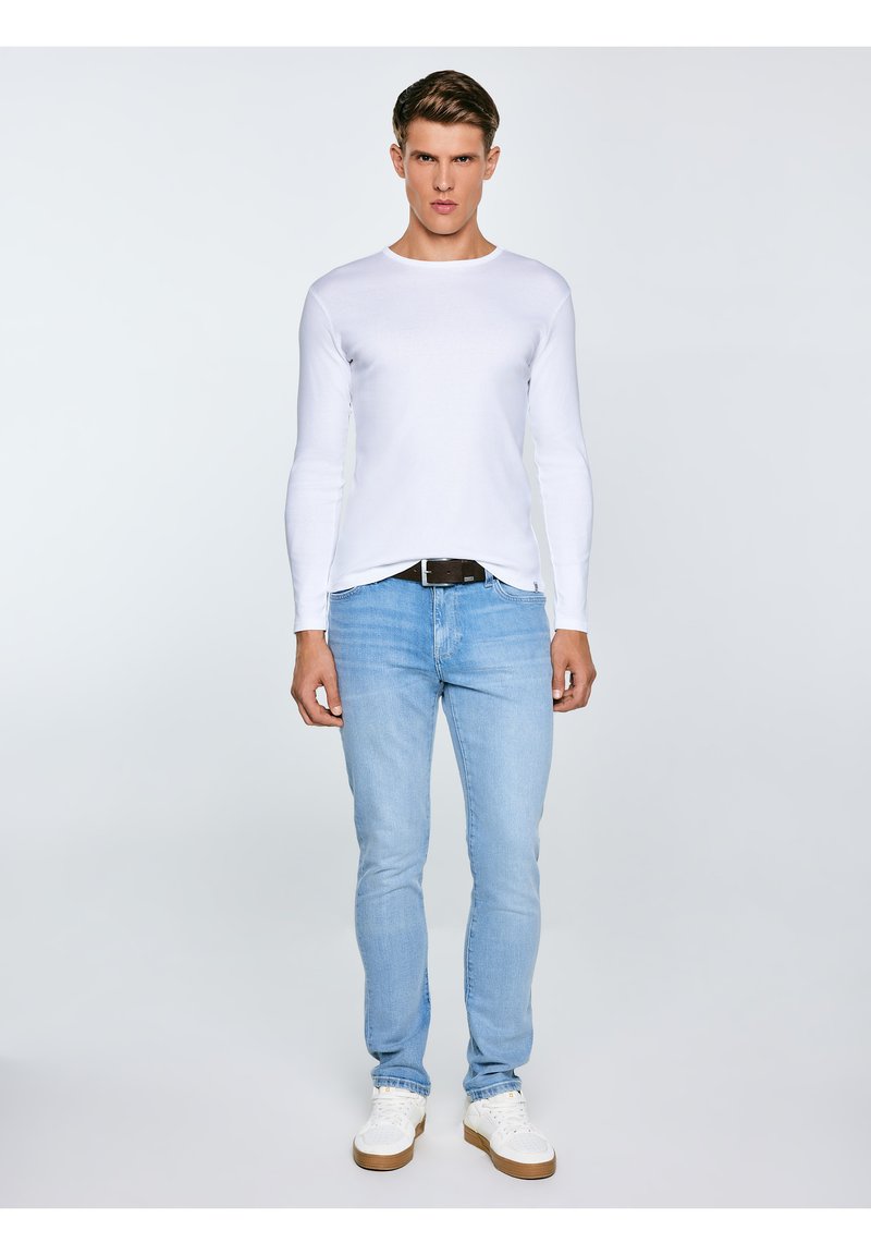 Jauns vīrietis stāv, valkājot baltu garroku kreklu, gaismazilas džinsas ar melnu jostu un baltas teniskas uz vienkrāsaina fona.