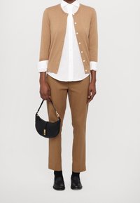 Beige pletený kardigan se zlatým logem, bílá košilová halenka, hnědé kalhoty, černá křížová taška se zlatými detaily a černé mokasíny.