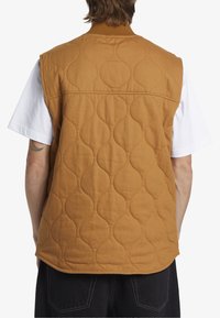 Prošívaná vesta v hnědé látce s ribbed límcem. Zadní strana má vlnkový vzor a zakřivený lem, obléká se přes bílé tričko.