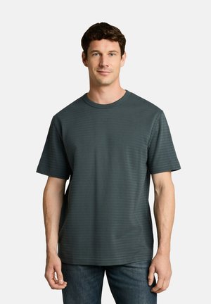 Mann mit kurzen braunen Haaren, der ein dunkelgrünes T-Shirt mit kurzen Ärmeln und Rundhalsausschnitt sowie blaue Jeans trägt und vor einem schlichten weißen Hintergrund steht.