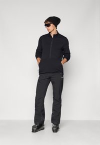 Giacca zip-up in pile nera con tasca frontale, abbinata a pantaloni impermeabili neri. Il modello indossa un berretto e occhiali da sole, mostrando uno stile da abbigliamento sportivo.
