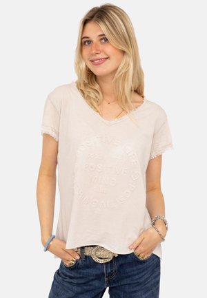 Young woman with blonde hair wears beige T-shirt with "Positive Life Positive Mind" text embossed, paired with blue jeans and accessories.