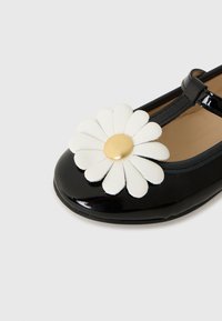 Schwarze Lackleder-Mary Jane-Schuh mit einer großen weißen Gänseblümchen-Dekoration mit gelbem Zentrum an der Spitze.