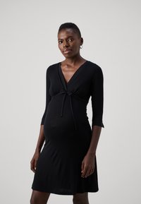 Zwarte zwangerschapsjurk met een V-hals, driekwartmouwen en een knoopdetail in de taille; zachte, rekbare stof met een vloeiende rok.