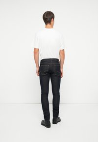 Jeans de denim azul oscuro, ajustados, con dos bolsillos traseros, costuras claras y una textura suave, combinados con una camiseta blanca lisa.