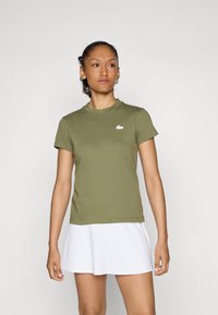 Olivenfarget T-skjorte med korte ermer, rund hals og en hvit Lacoste-logo på brystet. Stoffet ser jevnt og lett ut.