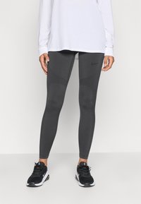 Sivé športové legíny s textúrovanými panelmi a logom Nike. Skombinované s dlhým bielym tričkom a čiernymi teniskami, ktoré ukazujú priliehavý dizajn.