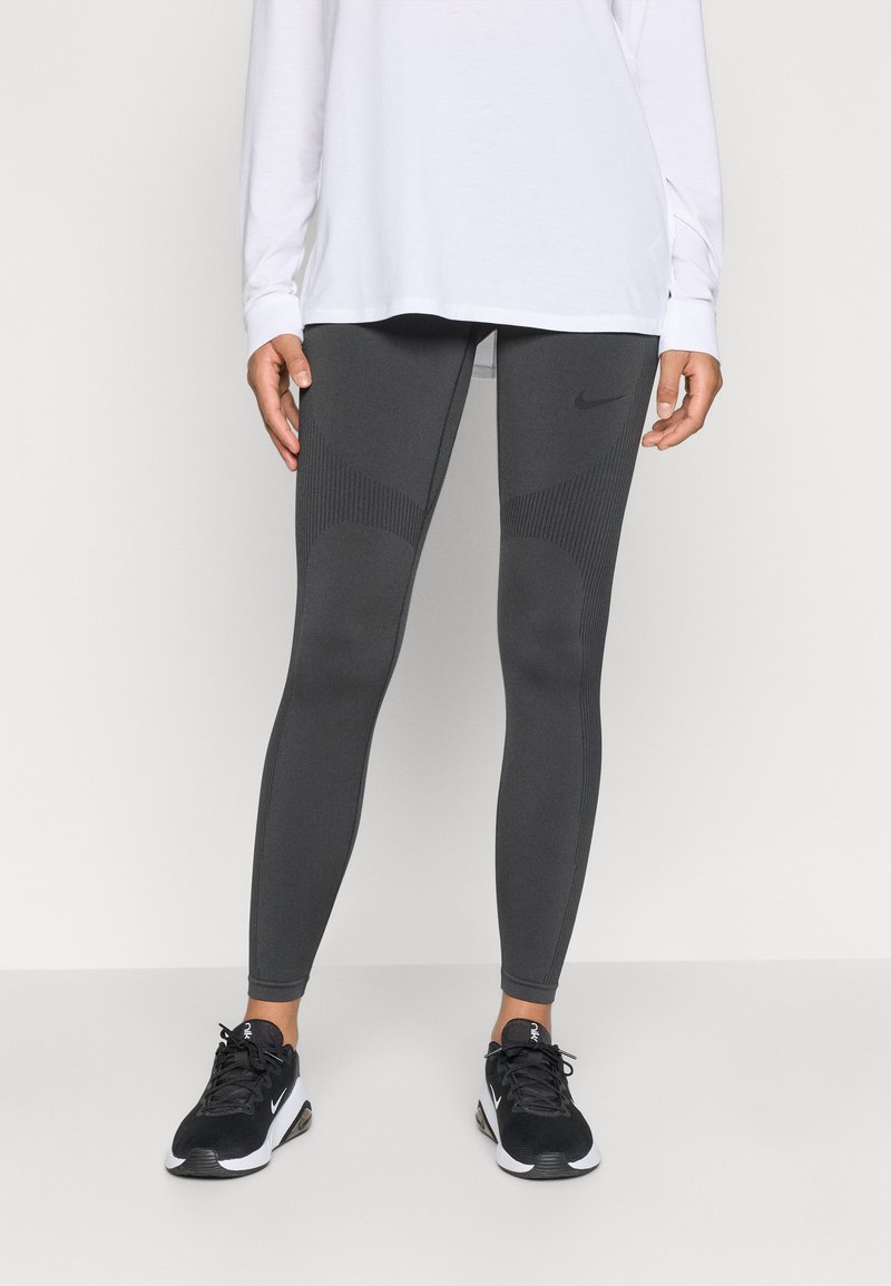 Sivé športové legíny s textúrovanými panelmi a logom Nike. Skombinované s dlhým bielym tričkom a čiernymi teniskami, ktoré ukazujú priliehavý dizajn.