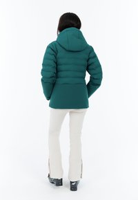 Tyrkysová péřová bunda s přiléhavým pasem, kapucí a prošívaným designem. V kombinaci se světle béžovými kalhotami a černými botami se zipy na kotnících.