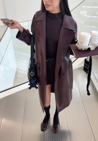 Trench-coat en cuir marron sur une robe noire, tenant un plateau avec des tasses de café. Porte-clés en peluche ours remarquable accroché à un sac noir.