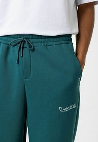 Teal katoenen joggers met een elastische tailleband met trekkoord, zijzakken en een wit geborduurd logo nabij de zoom. Gladde textuur en een ontspannen pasvorm.