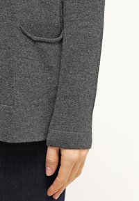 Szary dzianinowy sweter z widoczną kieszenią, ściągaczami na mankietach i miękką fakturą. Zbliżenie na rękaw i dłoń, podkreślające jego materiał i wzór.