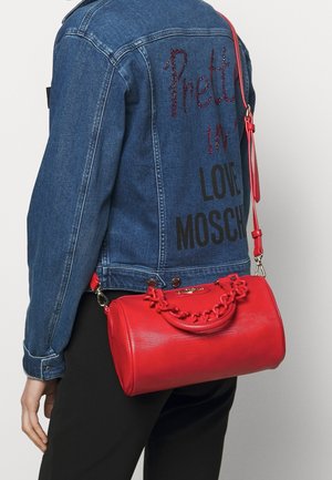 Osoba nosząca niebieską kurtkę dżinsową z napisem „Pretty in Love Moschino” i trzymająca czerwony walcowaty torebkę o fakturowanej powierzchni z łańcuszkowym detalem.