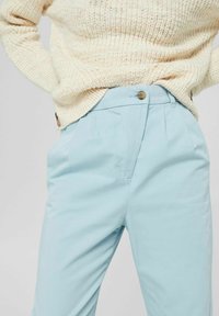 Pantalon bleu clair à texture lisse, avec un pli frontal, des poches latérales et une fermeture par bouton à la taille. Porté avec un pull crème.