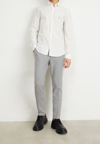 Hombre vestido con una camisa blanca de botones, pantalones grises claros y zapatos de vestir negros con cordones, de pie frente a una pared blanca lisa.